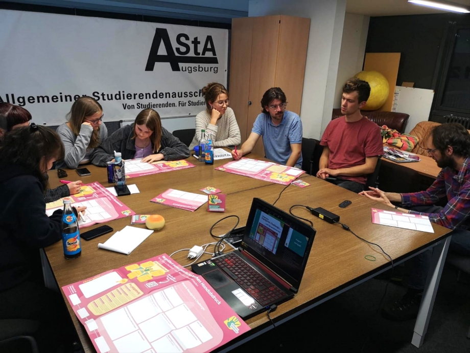 Der Seminarraum einer Hochschule mit 8 Personen an einem Tisch. Im Hintergrund das Banner des AStA der Uni Augsburg. 