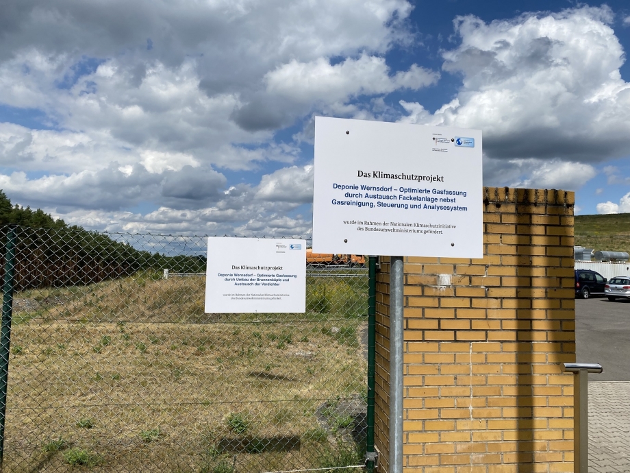 Schild am Zaun vor der Deponieanlage mit einer Beschreibung zum Projekt und den Logos der NKI und des BMWK