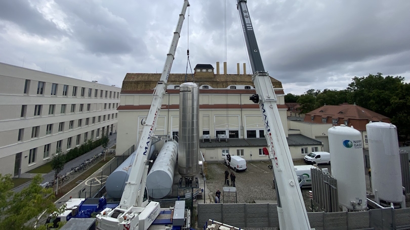 Zwei große Kräne heben zylindrische Objekte hinter zwei Gebäuden.