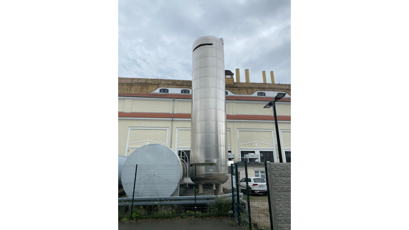 Ein grosser, zylindrischer Edelstahltank steht vor einem Industriegebäude mit mehreren Schornsteinen.