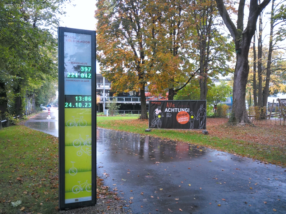 Eine Straße mit einer Säule, auf der die Anzahl der Fahrräder aufgeführt sind, im Hintergrund Herbstbäume und ein Gebäude