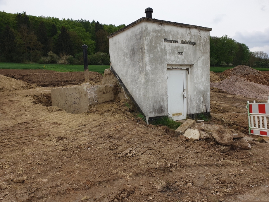 Alter Hochbehälter in Niederlistingen. Eine verputztes kastenförmiges Gebäude mit Eingangstür im Vordergrund. Darüber die Einschrift über der Tür "Wasserwerk Niederlistingen" und darunter das Erbauungsdatum "1933". Um die Anlage herum umgegrabene Erde der Baustelle, im Hintergrund eine Wiese, dahinter der Beginn eines Waldstücks.