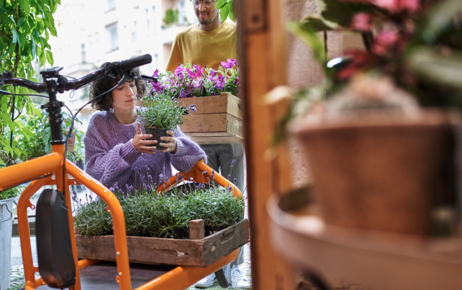 Die Inhaberin eines Blumenladens belädt ein orangenes E-Lastenfahrrad mit Pflanzen. Im Hintergrund steht ein Mitarbeiter, der eine Holzkiste mit Pflanzen bringt.