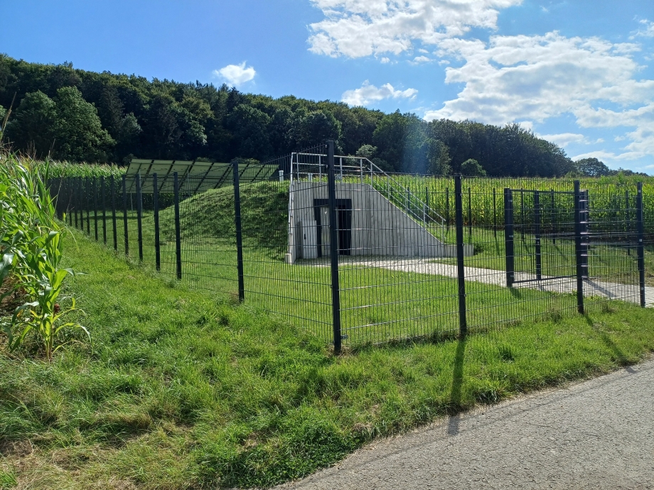 Seitenansicht der Pumpwerks in Niederlistingen. Die Anlage ist umzäunt mit einem schwarzen, hohen Metallzaun. Vor der Anlage eine Rasenfläche. Die Anlage besitzt im Vordergrund Betonwände mit der Eingangstür im Zentrum. Über den Betonwänden metallene Brüstungen. Hinter dem Eingang aus Beton eine hügelähnliche, von Gras bedeckte Erhöhung. Hinter der Anlage befinden sich mehrere Solarpanele, die auf der Wiese aufgestellt sind. Neben der Anlage Maisfelder und im Hintergrund der Beginn eines Waldstücks. 