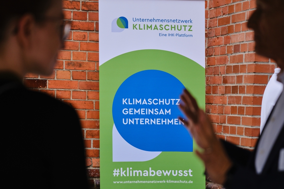 Zwei Personen im Gespräch vor einem Banner des Unternehmensnetzwerks Klimaschutz. Das Banner trägt die Aufschrift "Klimaschutz gemeinsam unternehmen" und "#klimabewusst" sowie die URL "www.unternehmensnetzwerk-klimaschutz.de".