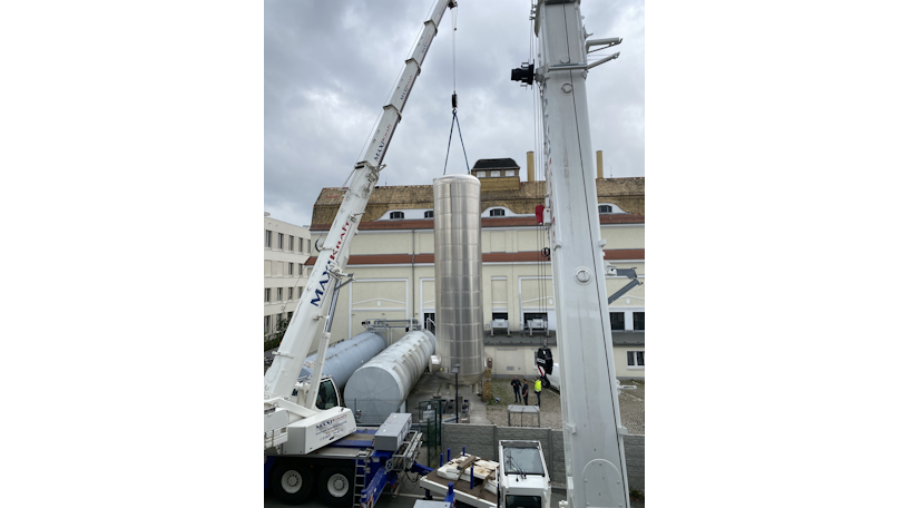 Ein großer zylindrischer Metallsilo wird von einem Kran über einer Baustelle gehoben. Darunter überwachen mehrere Personen in Sicherheitsausrüstung den Vorgang. Im Hintergrund sind Gebäude zu sehen.