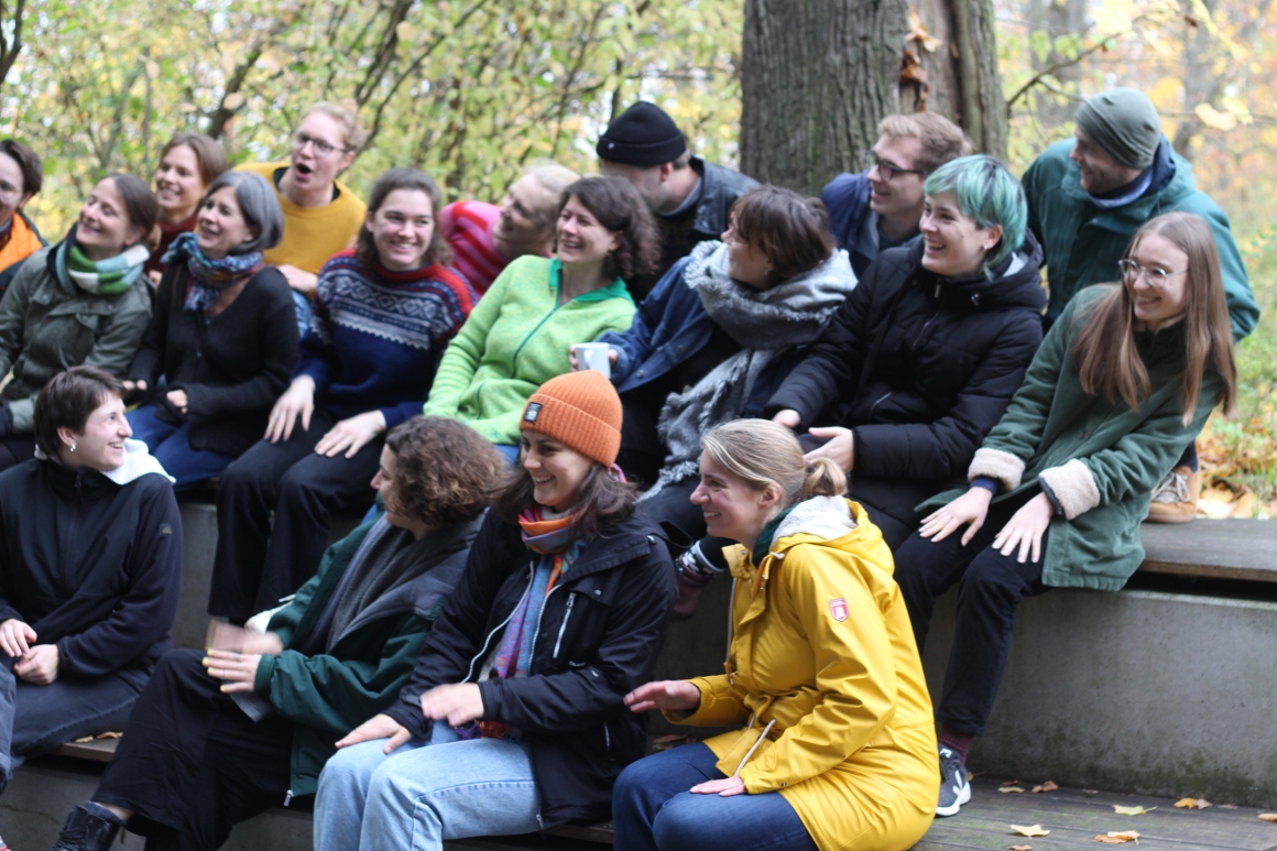 Ein Foto von 18 Personen in einem Park. Sie sitzen in drei Reihen auf einer Treppe. Dabei lachen sie ausgelassen. 