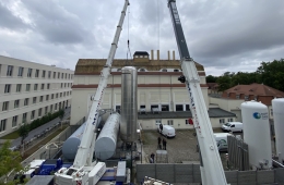 Zwei große Kräne heben zylindrische Objekte hinter zwei Gebäuden.