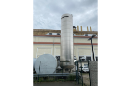 Ein grosser, zylindrischer Edelstahltank steht vor einem Industriegebäude mit mehreren Schornsteinen.