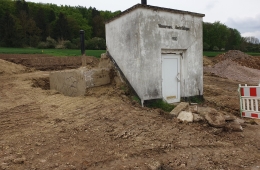 Alter Hochbehälter in Niederlistingen. Eine verputztes kastenförmiges Gebäude mit Eingangstür im Vordergrund. Darüber die Einschrift über der Tür "Wasserwerk Niederlistingen" und darunter das Erbauungsdatum "1933". Um die Anlage herum umgegrabene Erde der Baustelle, im Hintergrund eine Wiese, dahinter der Beginn eines Waldstücks.