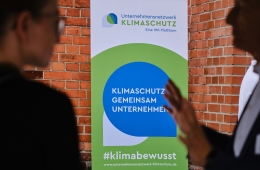 Zwei Personen im Gespräch vor einem Banner des Unternehmensnetzwerks Klimaschutz. Das Banner trägt die Aufschrift "Klimaschutz gemeinsam unternehmen" und "#klimabewusst" sowie die URL "www.unternehmensnetzwerk-klimaschutz.de".