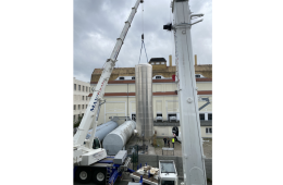 Ein großer zylindrischer Metallsilo wird von einem Kran über einer Baustelle gehoben. Darunter überwachen mehrere Personen in Sicherheitsausrüstung den Vorgang. Im Hintergrund sind Gebäude zu sehen.