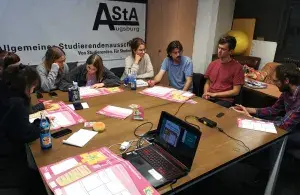 Der Seminarraum einer Hochschule mit 8 Personen an einem Tisch. Im Hintergrund das Banner des AStA der Uni Augsburg. 