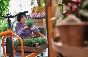 Die Inhaberin eines Blumenladens belädt ein orangenes E-Lastenfahrrad mit Pflanzen. Im Hintergrund steht ein Mitarbeiter, der eine Holzkiste mit Pflanzen bringt.