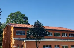 Orangefarbene Turnhalle mit mehreren Fenstern, im Vordergrund und im Hintergrund sind zwei Bäume.