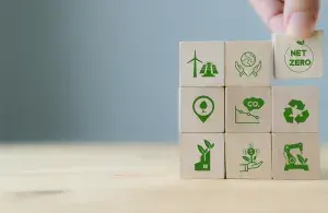 Holzbausteine mit grünen Klimaschutzsymbolen, wie eine Pyramide angeordnet, der oberste Baustein mit der Aufschrift 'net Zero' wird von einer Hand auf die Spitze gesetzt. 