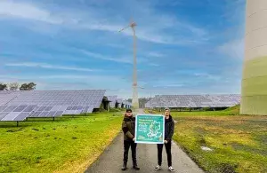 Zwei Personen stehen vor Windrädern und Photovoltaikanlagen und halten ein Plakat mit dem Keyvisual von #StudyGreenEnergy